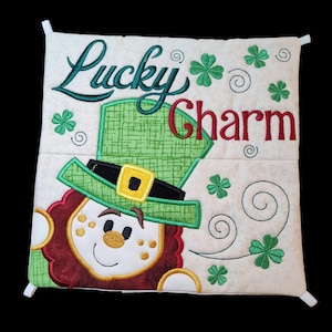 May include: A white pillow with a green, red, and yellow embroidered leprechaun wearing a green hat with a gold buckle. The pillow has the text "Lucky Charm" embroidered on it.