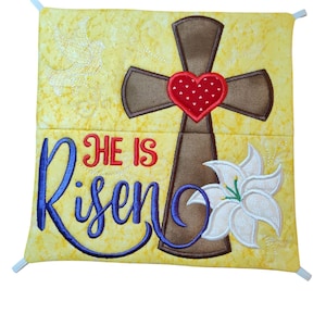 May include: A yellow fabric square with a brown cross, a red heart with white polka dots, a white lily, and the text "He is Risen" in blue lettering.