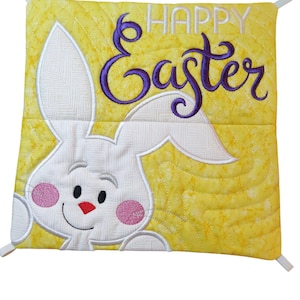 May include: A yellow quilted pillow with a white bunny face and the words "Happy Easter" embroidered in purple.