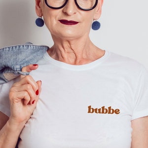May include: A white t-shirt with the word "bubbe" printed in brown letters on the front. The shirt is being worn by a person with short gray hair and blue earrings.