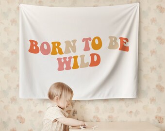 May include: A white tapestry with the text "BORN TO BE WILD" in colourful, retro-style letters. A young child is standing in front of the tapestry, looking at a small wooden table.