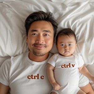 以下が含まれることがあります： 白いTシャツを着た父親と息子。Tシャツには茶色の文字で「ctrl c」と「ctrl v」と印刷されています。白いベッドに横になってカメラを見ています。