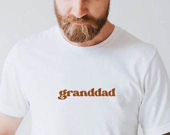 May include: White t-shirt with the word "granddad" in a brown retro font. The shirt is made of a soft material and is a casual style. The text is centered on the chest.