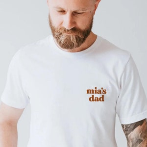 May include: White t-shirt with a brown text design that says "mia's dad" on the front.