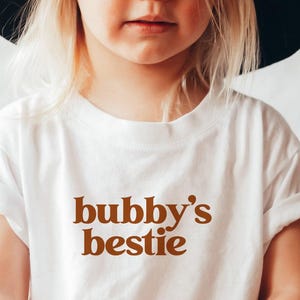 May include: White toddler t-shirt with brown text that reads "bubby's bestie".