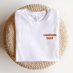 May include: White t-shirt with the text "custom text" in brown lettering on the front.