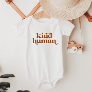 Peut inclure: Combinaison b&eacute;b&eacute; blanche avec un texte brun qui dit "kind human" dans une police r&eacute;tro.