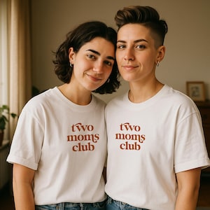 May include: Two people wearing off-white t-shirts with the text "Two Moms Club" in a brown, retro font. The shirts are paired with blue jeans. The background is a neutral-toned room with a window and a wooden cabinet.