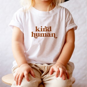 Peut inclure: Un t-shirt blanc avec le texte "kind human" imprim&eacute; en marron sur le devant. Le t-shirt est port&eacute; par un enfant.