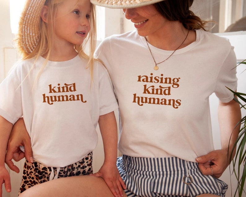 Peut inclure: Un t-shirt blanc avec le texte "raising kind humans" en lettres brunes. Un enfant porte un t-shirt blanc avec le texte "kind human" en lettres brunes. L'enfant porte une jupe &agrave; imprim&eacute; l&eacute;opard brun et blanc. L'adulte porte une jupe ray&eacute;e bleu et blanc.