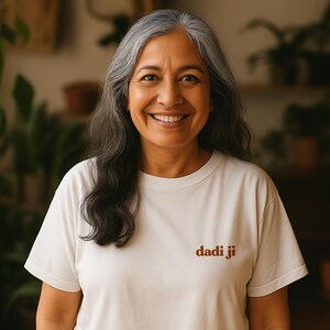 May include: A person with long grey hair smiles, wearing a cream t-shirt. The t-shirt features the brown text "dadi ji". The background is blurred, showing plants and a warm, natural light.