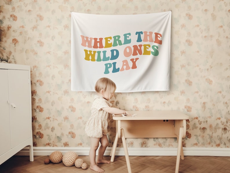 Pu&ograve; includere: Un arazzo bianco con il testo "Where the Wild Ones Play" in lettere colorate in stile retr&ograve;. L'arazzo &egrave; appeso a una parete con carta da parati floreale. Un bambino piccolo &egrave; in piedi davanti a un tavolo di legno.
