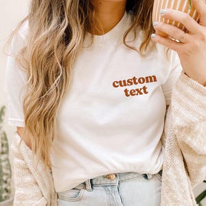 May include: White t-shirt with the text "custom text" in brown lettering.