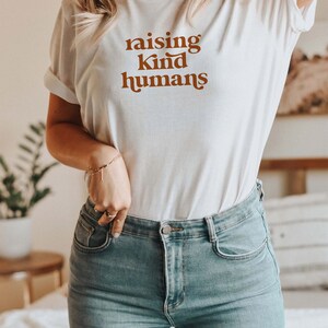 Peut inclure: Un t-shirt blanc avec le texte "raising kind humans" imprim&eacute; en marron sur le devant. Le t-shirt est port&eacute; par une personne aux cheveux blonds et un jean bleu.