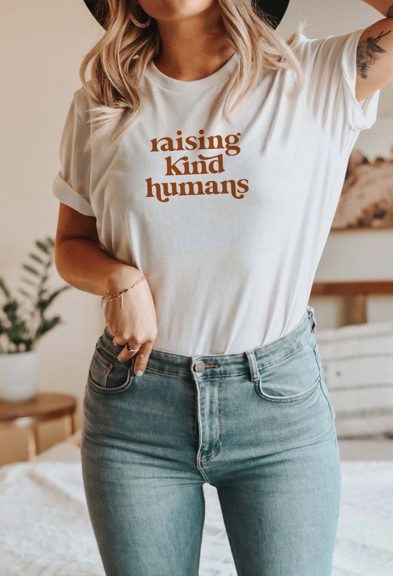 Peut inclure: Un t-shirt blanc avec le texte "raising kind humans" imprim&eacute; en marron sur le devant. Le t-shirt est port&eacute; par une personne aux cheveux blonds et un jean bleu.