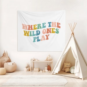 Pu&ograve; includere: Un arazzo bianco con il testo "Where the Wild Ones Play" in lettere colorate in stile retr&ograve;. L'arazzo &egrave; appeso a una parete bianca in una stanza con un teepee, un peluche e uno scaffale di legno con giocattoli.