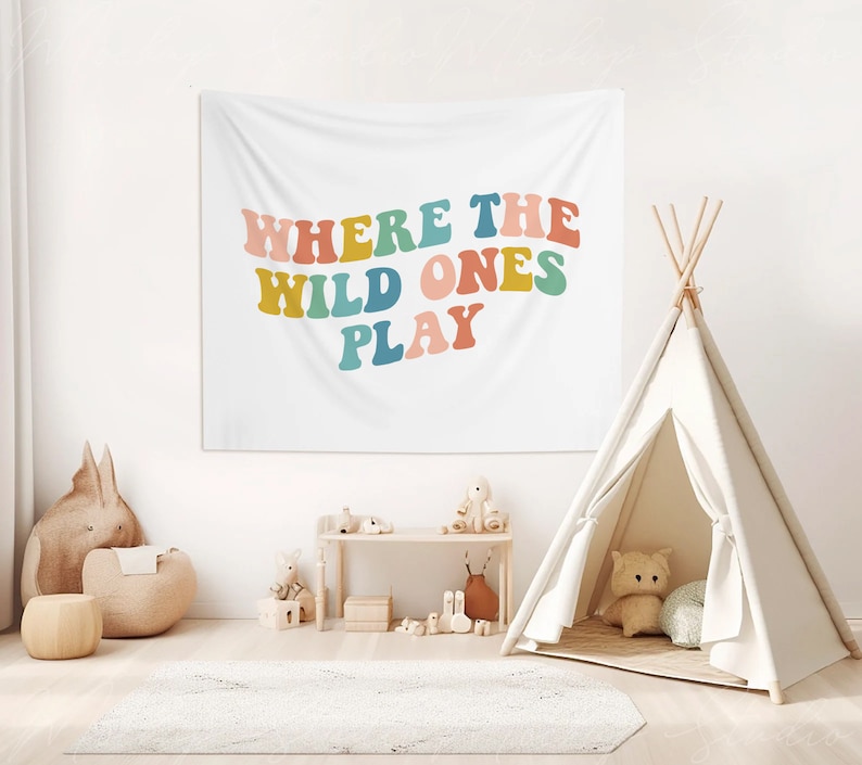 Pu&ograve; includere: Un arazzo bianco con il testo "Where the Wild Ones Play" in lettere colorate in stile retr&ograve;. L'arazzo &egrave; appeso a una parete bianca in una stanza con un teepee, un peluche e uno scaffale di legno con giocattoli.
