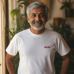 May include: A white t-shirt with the word "dada" in brown lettering. The short-sleeved shirt is made of a lightweight material. The person wearing the shirt is smiling, with plants and a window in the background.