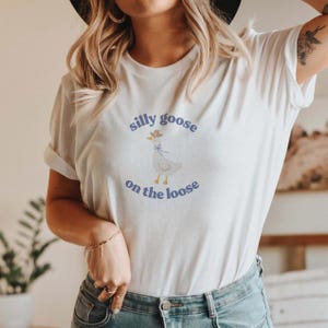 May include: White t-shirt with a blue and white illustration of a goose wearing a cowboy hat and the text "silly goose on the loose".