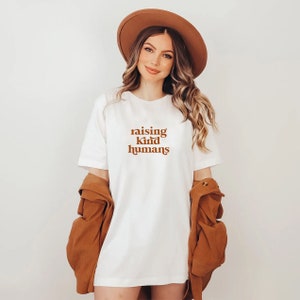 Peut inclure: T-shirt blanc avec un graphisme brun qui dit "raising kind humans".