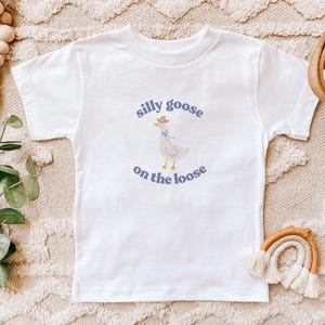 May include: White t-shirt with a blue and white illustration of a goose wearing a cowboy hat. The text "silly goose on the loose" is printed in blue below the goose.