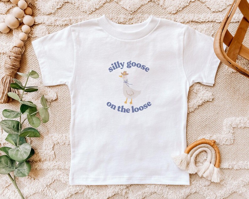 May include: White t-shirt with a blue and white illustration of a goose wearing a cowboy hat. The text "silly goose on the loose" is printed in blue below the goose.