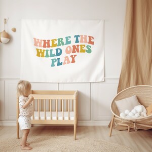 Pu&ograve; includere: Un arazzo bianco con il testo "Where the Wild Ones Play" in lettere colorate in stile retr&ograve;. L'arazzo &egrave; appeso a una parete bianca in una cameretta con una culla di legno e una sedia in vimini.