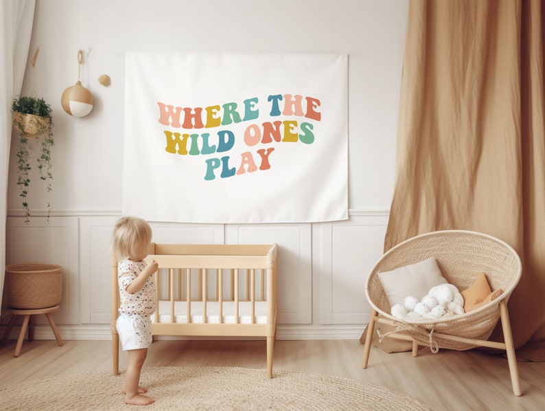 Pu&ograve; includere: Un arazzo bianco con il testo "Where the Wild Ones Play" in lettere colorate in stile retr&ograve;. L'arazzo &egrave; appeso a una parete bianca in una cameretta con una culla di legno e una sedia in vimini.