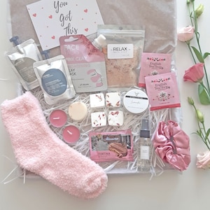 May include: A pink and white gift box filled with self-care items, including a pink face mask, bath salts, bath bombs, candles, a pair of fluffy socks, and a hair scrunchie. The box also includes a small bag of tea and a chocolate bar.