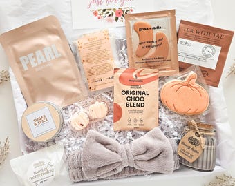 May include: A gift box filled with self-care items. Includes a headband, eye masks, tea, a pumpkin-shaped cookie, a candle, a bag of face mask, and a card that says "just for you". The box is lined with white paper.