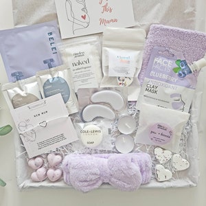 May include: A gift box filled with self-care items for a new mother. The box contains a face mask, eye patches, soap, candles, bath salts, a headband, and heart-shaped wax melts. The box also includes a card with the text "New Mum" and "This Mama".
