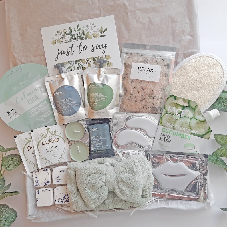 May include: A gift basket filled with self-care items, including face masks, bath salts, eye masks, and candles. The basket is decorated with green foliage and the words "just to say" on a card.
