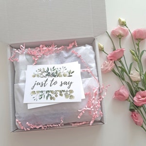 May include: A white gift box filled with pink tissue paper and a card that says "just to say" with a green floral design.