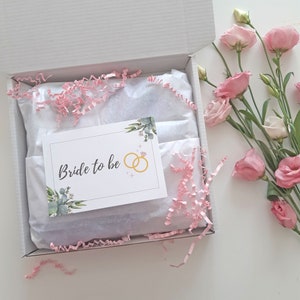 May include: A white gift box with pink tissue paper and a card that says "Bride to be" with a gold ring illustration. The box is open and there are pink flowers in the background.