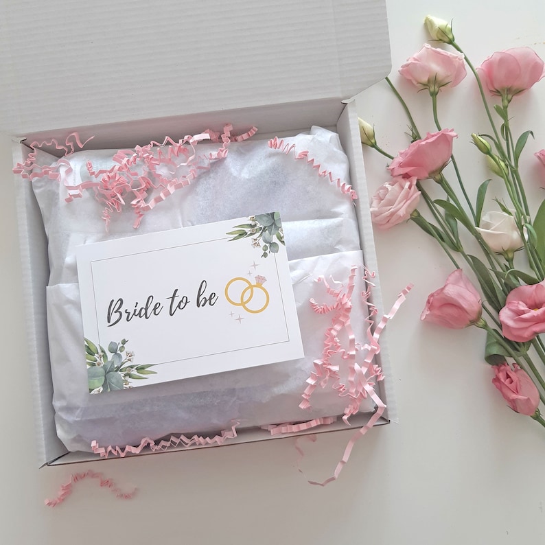 May include: A white gift box with pink tissue paper and a card that says "Bride to be" with a gold ring illustration. The box is open and there are pink flowers in the background.