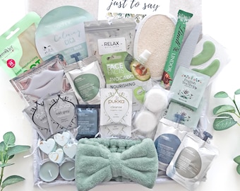 May include: A gift box containing a sage green plush headband, various skincare sachets including face masks and bath salts, two white bath bombs, a round loofah, a Lindt chocolate stick, English Tea Shop tea bags, and a card reading just to say.