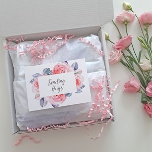 May include: A white gift box with pink tissue paper and a card that says "Sending Hugs" with a floral design. The box is open and there are pink flowers on the table next to it.
