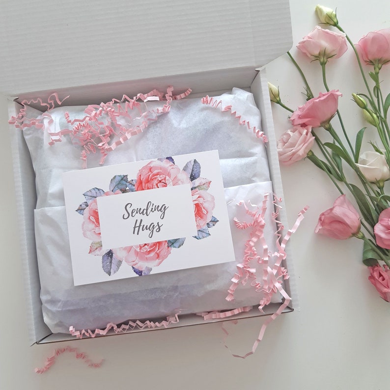 May include: A white gift box with pink tissue paper and a card that says "Sending Hugs" with a floral design. The box is open and there are pink flowers on the table next to it.
