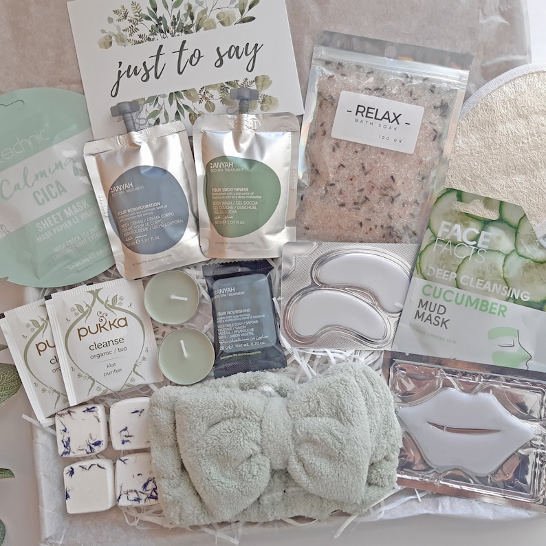 May include: A green and white gift box filled with self-care items, including bath salts, face masks, and candles. The box also includes a green headband with a bow, a small bag of tea, and a card that says "just to say".