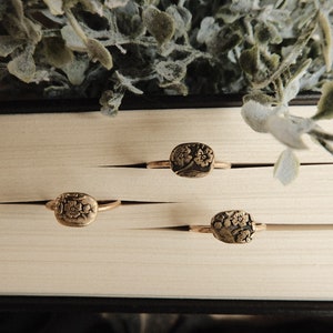 Puede incluir: Tres anillos de oro con diseños florales sobre una superficie blanca. Los anillos están dispuestos en fila, con el anillo más grande en la parte superior y el anillo más pequeño en la parte inferior.