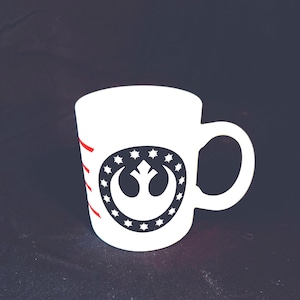 May include: White ceramic mug featuring a black and white Rebel Alliance symbol from Star Wars. The symbol is encircled by a ring of white stars. The mug has red stripes on the side.