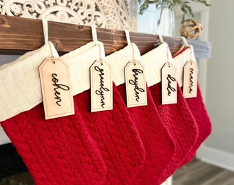 May include: Five red knitted stockings with white cuffs hang from a wooden mantelpiece. Each stocking has a wooden tag with a name written in black calligraphy: "mama", "dala", "brayden", "gracelyn", and "cohen".