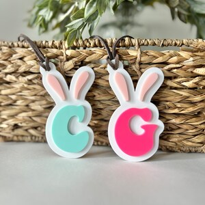 May include: Two white bunny-shaped ornaments with pink ears. The bunny on the left has a light blue letter "C" and the bunny on the right has a pink letter "G".