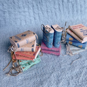 Eight pairs of earrings in the shape of miniature books in a pile. They each have a different cover and different coloured hooks.