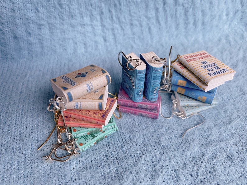 Eight pairs of earrings in the shape of miniature books in a pile. They each have a different cover and different coloured hooks.