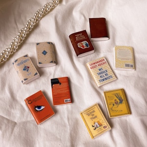 Five pairs of miniature books laid out flat to show the cover. These are all yellow orange or brown in colour. Les miserables. Handbook for the recently deceased. Clockwork orange. We should all be feminists. Winnie the pooh.