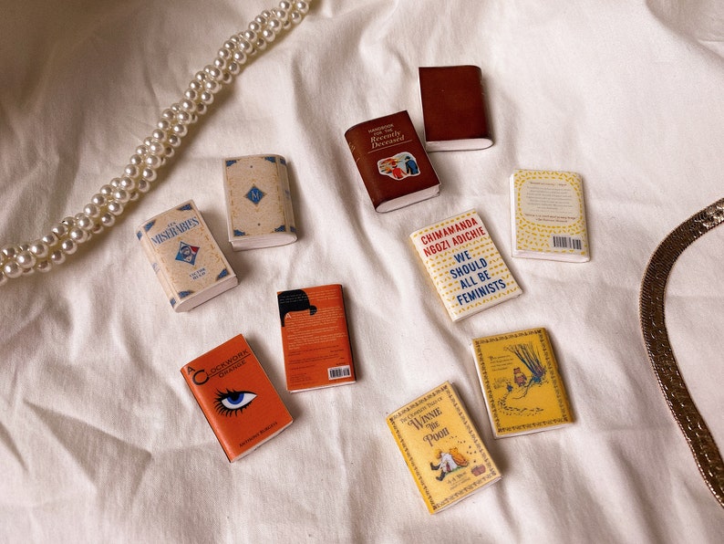 Five pairs of miniature books laid out flat to show the cover. These are all yellow orange or brown in colour. Les miserables. Handbook for the recently deceased. Clockwork orange. We should all be feminists. Winnie the pooh.