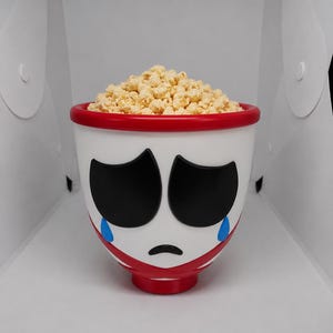 May include: A white bowl with a red rim and base, featuring a stylized sad face with large black eyes, a small black mouth, and two blue teardrops. The bowl is filled with a mound of light yellow, textured, popcorn-like snack pieces.