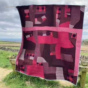 May include: A patchwork quilt with a geometric design in shades of pink, red, and brown. The quilt is made of different fabrics, including plaid and gingham.