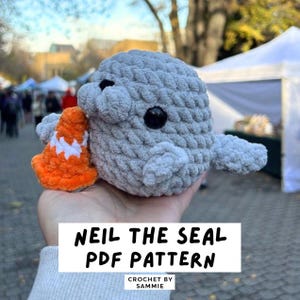 May include: A gray crocheted seal plush toy with black plastic eyes, holding a small orange and white crocheted traffic cone. The image includes the text NEIL THE SEAL PDF PATTERN and CROCHET BY SAMMIE in black lettering on a white background.